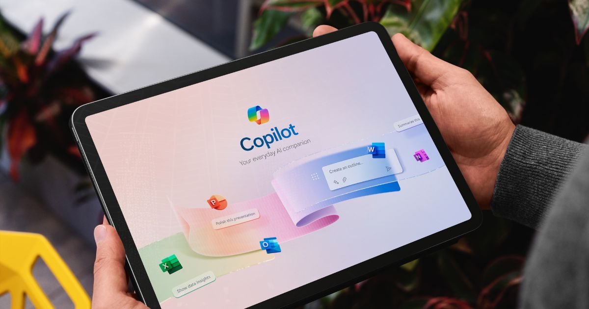 A imagem mostra um tablet sendo segurado com ambas as mãos, exibindo a interface do Microsoft Copilot com o texto “Copilot – Your everyday AI companion”. A tela apresenta um design limpo e moderno, com fundo claro e elementos gráficos coloridos que representam aplicativos do pacote Microsoft 365, como Word, Excel, PowerPoint e Outlook, sugerindo integração e produtividade. O cenário ao redor inclui detalhes de um ambiente interno com plantas e uma cadeira amarela parcialmente visível, transmitindo um clima contemporâneo e descontraído. A iluminação é natural, destacando bem o dispositivo e a interface.