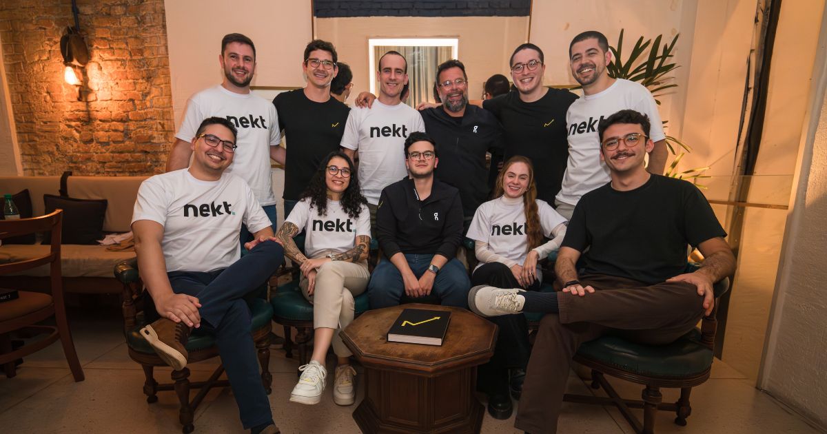 A foto mostra um grupo de dez pessoas reunidas em um ambiente interno com decoração acolhedora. O espaço tem paredes de tijolos aparentes, iluminação quente e móveis de madeira. No centro, há uma mesa baixa com um caderno preto que possui um símbolo em forma de “V” amarelo na capa. As pessoas estão posicionadas em duas fileiras: algumas sentadas em poltronas e outras em pé atrás delas. Várias pessoas vestem camisetas brancas com a palavra “nekt” impressa na frente, enquanto outras usam roupas escuras, incluindo camisetas pretas com um pequeno logotipo no lado esquerdo do peito. Ao fundo, há uma planta decorativa e detalhes como uma luminária de parede à esquerda.