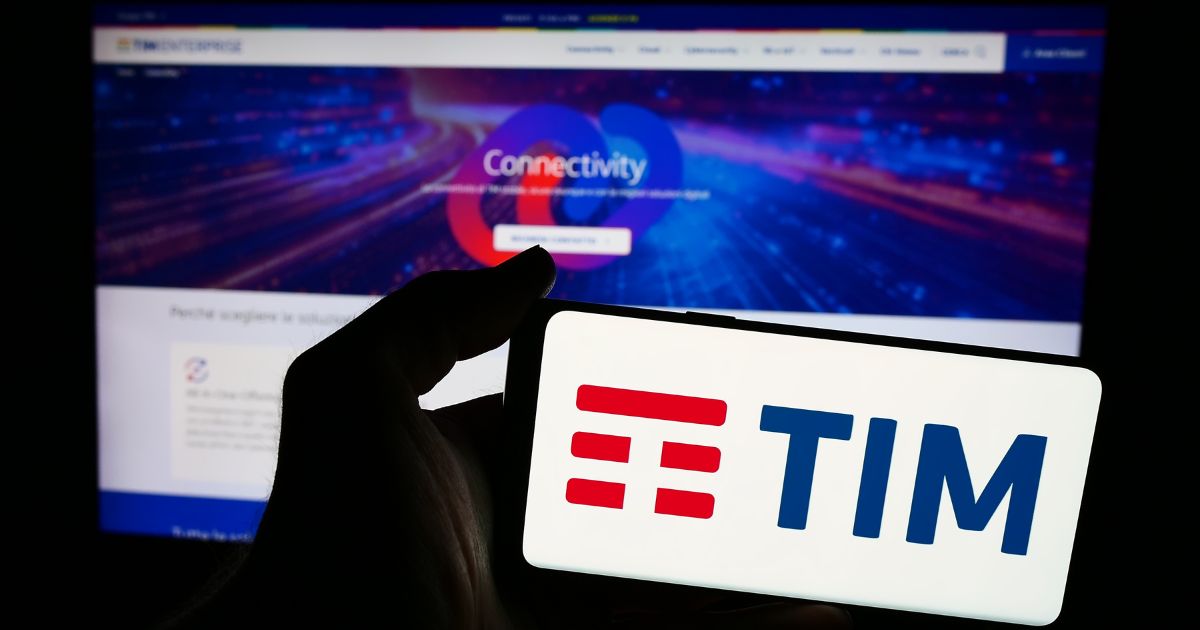 A imagem mostra um dispositivo móvel sendo segurado por uma mão, exibindo o logotipo da TIM em destaque, com letras azuis e três linhas vermelhas estilizadas à esquerda. Ao fundo, há uma tela de computador desfocada mostrando um site corporativo com a palavra “Connectivity” em evidência, acompanhada de elementos gráficos coloridos que remetem a tecnologia e redes digitais. A cena sugere um contexto relacionado a telecomunicações, conectividade ou serviços digitais, com iluminação que destaca o logotipo no primeiro plano.