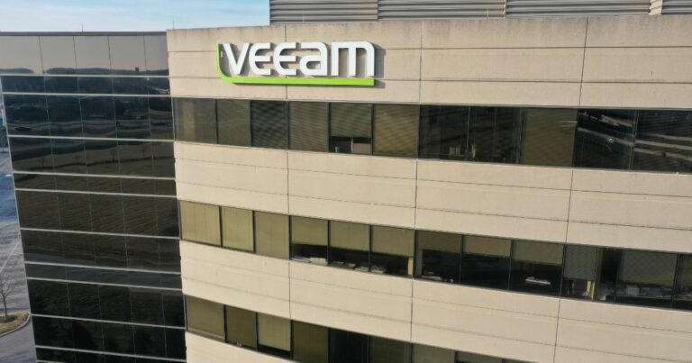 Um prédio comercial moderno aparece em destaque, com fachada composta por painéis de vidro escuro e áreas de concreto claro. Na parte superior da construção, há um letreiro grande com o nome “Veeam” em letras brancas e um detalhe verde sob a marca. As janelas refletem o ambiente externo e revelam parte do interior dos andares. A cena foi capturada de um ângulo levemente elevado, mostrando boa parte da lateral do edifício.