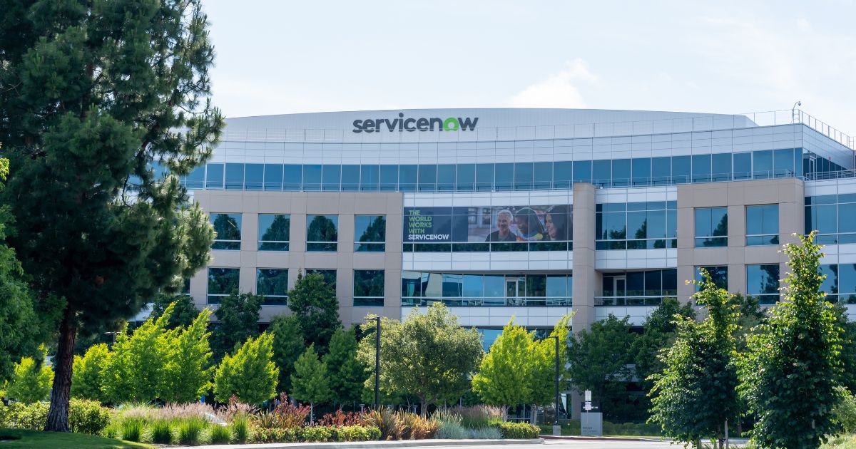 A imagem mostra a fachada de um edifício corporativo moderno com o logotipo ServiceNow no topo, em letras verdes e cinzas. A construção possui grandes janelas espelhadas e uma estrutura arquitetônica com linhas retas e tons claros, transmitindo um design contemporâneo. Em frente ao prédio, há uma área bem cuidada com árvores altas, arbustos e gramado, sugerindo um ambiente agradável e sustentável. O céu está claro, indicando um dia ensolarado, e a iluminação natural destaca a vegetação e a fachada do edifício.