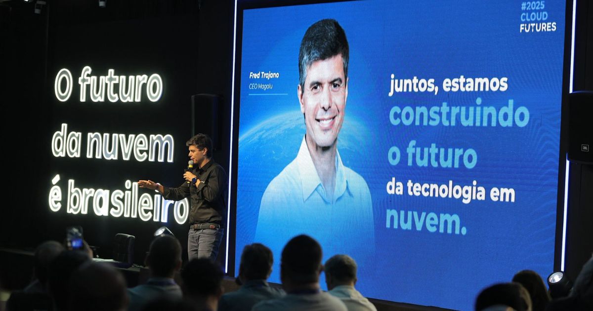 A imagem mostra um evento corporativo ou conferência com foco em tecnologia e computação em nuvem. No lado esquerdo, há um grande painel preto com a frase em destaque: “O futuro da nuvem é brasileiro” À direita, um painel azul exibe o texto: “juntos, estamos construindo o futuro da tecnologia em nuvem.” No canto superior direito do painel azul, aparece a marcação #2025 CLOUD FUTURES, indicando o tema ou nome do evento. Há também uma imagem de uma pessoa no painel, mas não será mencionada conforme solicitado. Em primeiro plano, uma pessoa está apresentando com microfone, voltada para o público, que está sentado e acompanhando a apresentação. O ambiente é moderno, com iluminação voltada para os painéis, reforçando a atmosfera tecnológica e inovadora. (Magalu)