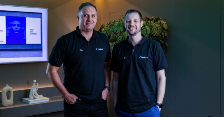 A imagem mostra duas pessoas vestindo camisetas pretas com o logotipo Navita no lado esquerdo do peito, indicando que estão representando a empresa. Ambas estão em pé, lado a lado, em um ambiente interno moderno e bem iluminado. No fundo, há uma parede com uma tela exibindo um slide com tons de azul e branco, contendo o texto “TECH FOR BUSINESS”, sugerindo um contexto corporativo ou tecnológico, possivelmente relacionado a uma apresentação ou reunião. À esquerda, sobre um móvel, há objetos decorativos, incluindo uma escultura clara e um vaso, enquanto à direita há uma planta verde que adiciona um toque natural ao espaço. (Nava)